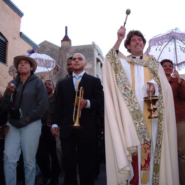 Man with trumpet next to priest blessing crowd in parade down street. 