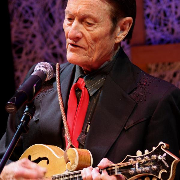 An older man with dark hair, black shirt and coat, and a red tie sings into a microphone and plays the mandolin 
