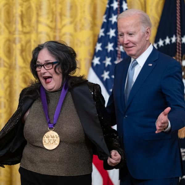 Older white male in blue suit posing with Hispanic woman in glasses wearing a black jacket in front of flags and gold curtain.