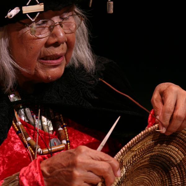 Native woman with white hair wearing headdress and holding up basket. 