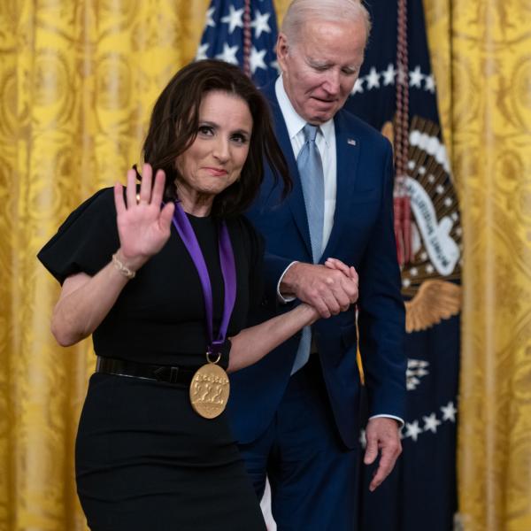 Older white male in blue suit posing with white woman in black dress in front of flags and gold curtain.