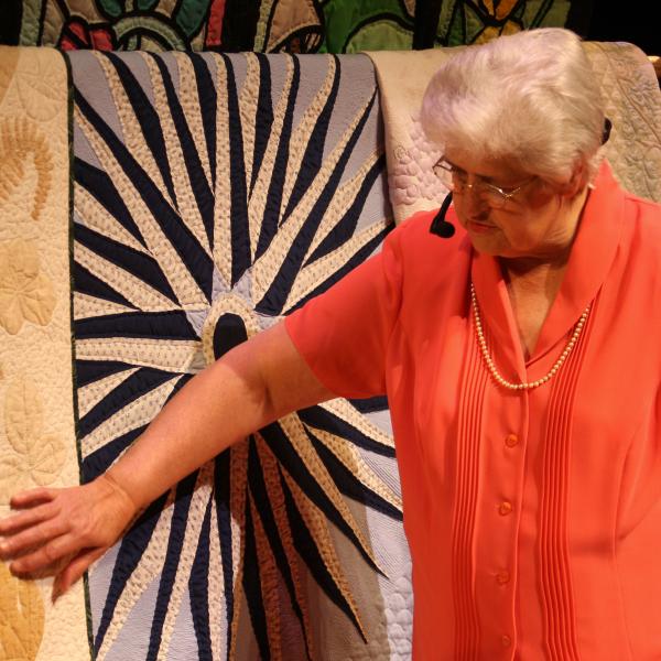 White woman in an orange dress standing in front of three quilts hung up behind her. 