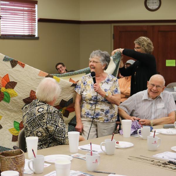An older white woman stands at a table with a microphone in front of her handmade quilt