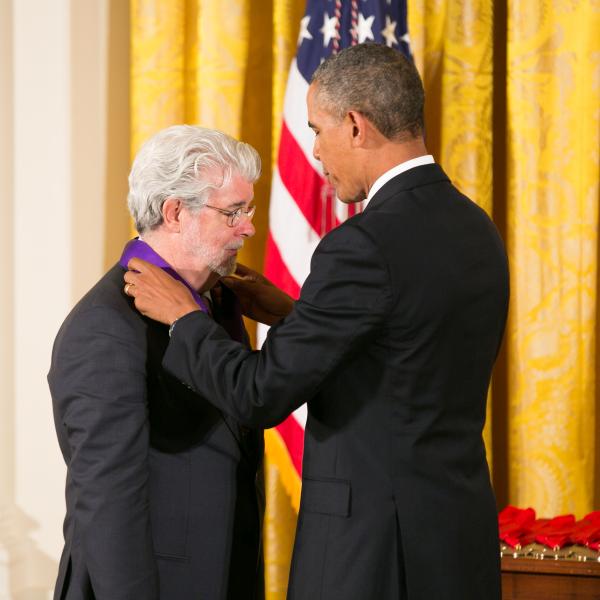 Man with gray hair and gray beard receiving a medal from tall black man in a suite in front of an American flag. 