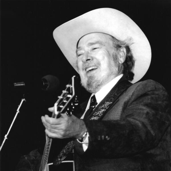 Man with gray beard in white hat playing a guitar. 