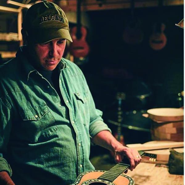 Man wearing a baseball cap and blue denim shirt looking down at a guitar. 