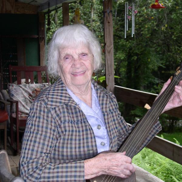 Elderly woman with gray hair sitting on porch playing a banjo. 