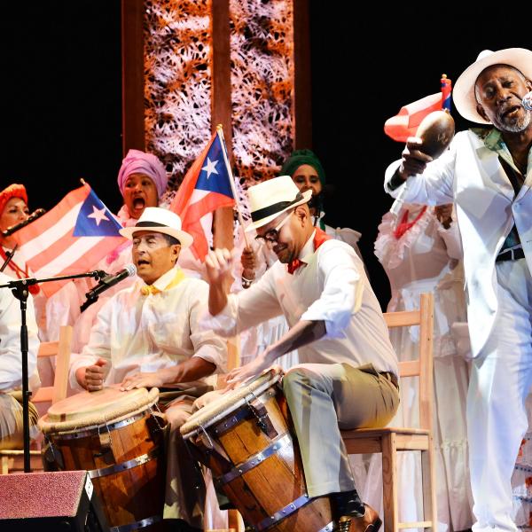 On stage: Puerto Rican man (right) wears a white hat and suit performing on stage alongside seven musicians (three men are playing drums and four women are behind them holding Puerto Rico flags).