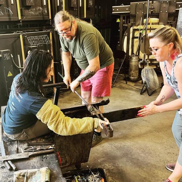 A group of three people uses metal tools to refine a piece of glass-blown art.