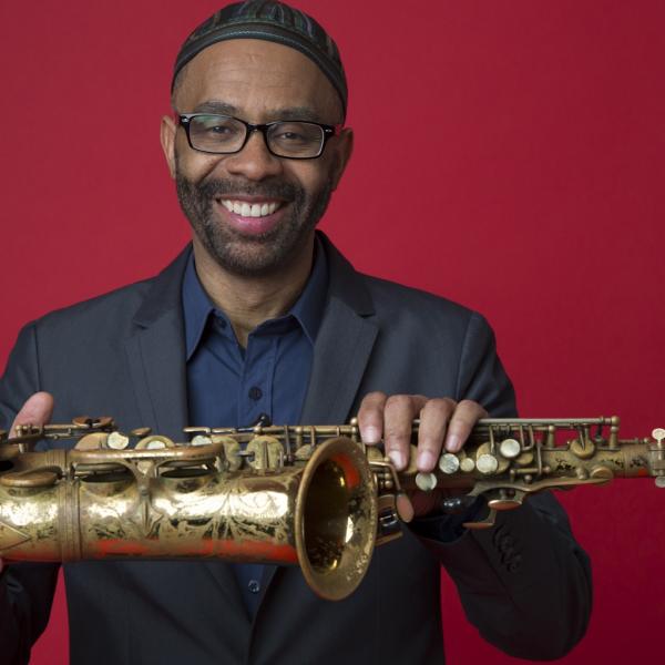 Black man wearing glasses and skullcap in blue shirt holding saxophone sideways in front of red background.