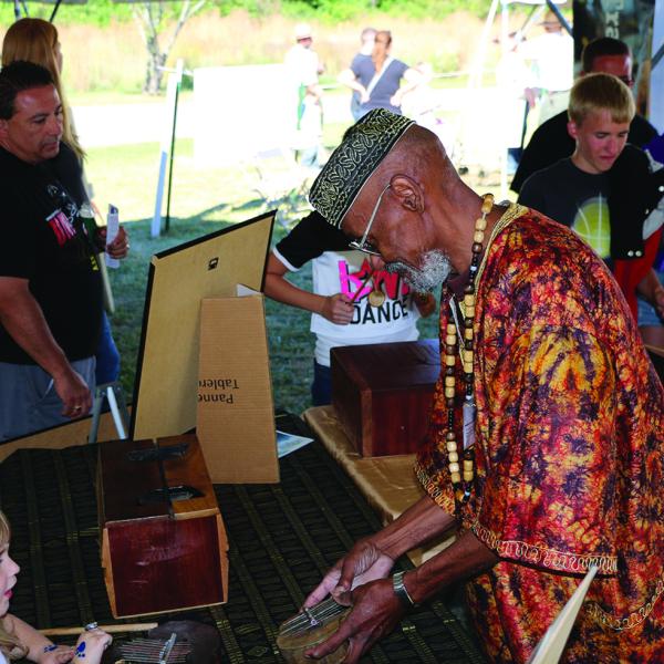 An older man shows a small drum to a young girl while in the background other people look at handmade drums