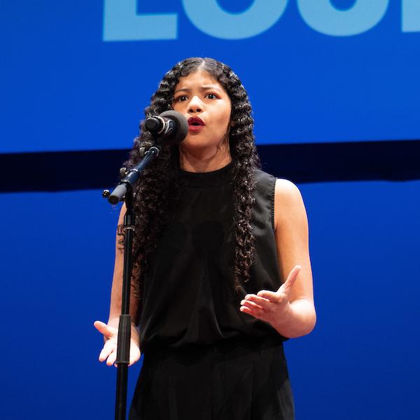 A young woman gestures with her hands while standing behind a microphone.