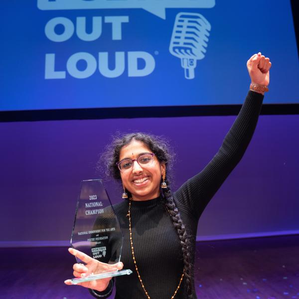 A young woman with two long braids holds a trophy in one hand with her other hand in the air. Behind her is a screen with the Poetry Out Loud logo.