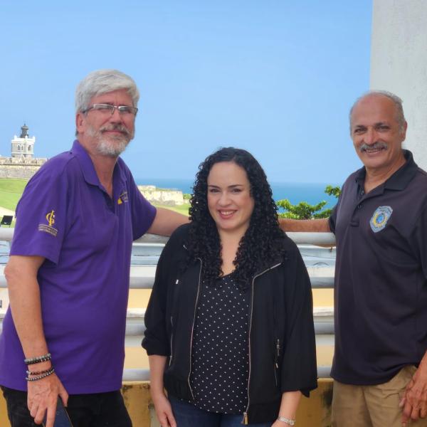 A man with gray hair and goatee, wearing glasses and a blue shirt; woman with long dark hair wearing a black shirt; bald man with moustache where dark blue shirt. 