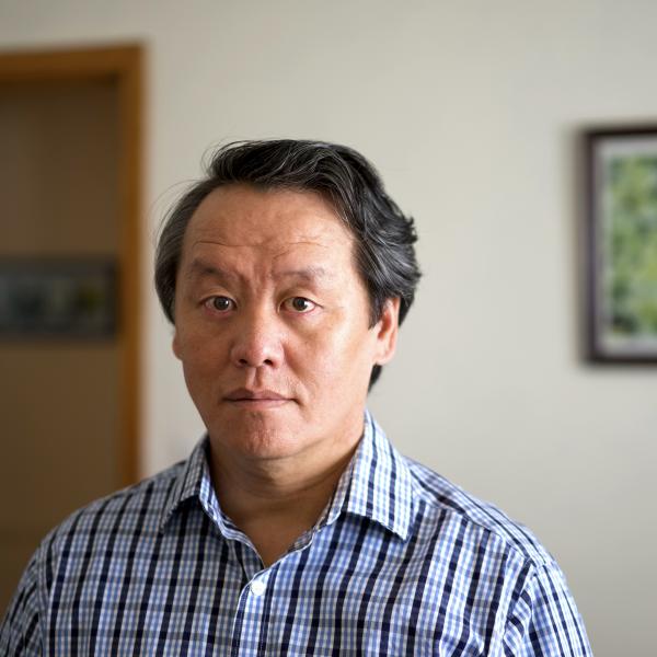 Portrait of Asian man in blue striped shirt with a door and a painting in the background.