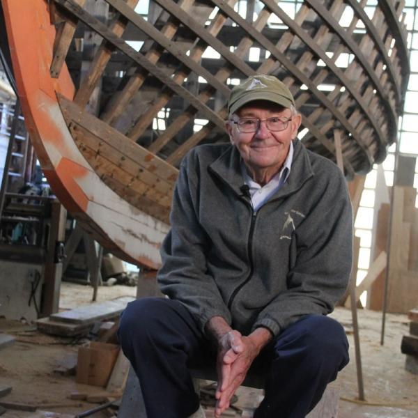 Man sits on a stool in front of the frame of a wooden boat during construction