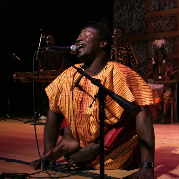Black man in African shirt and disabled legs singing into a mic on stage. 