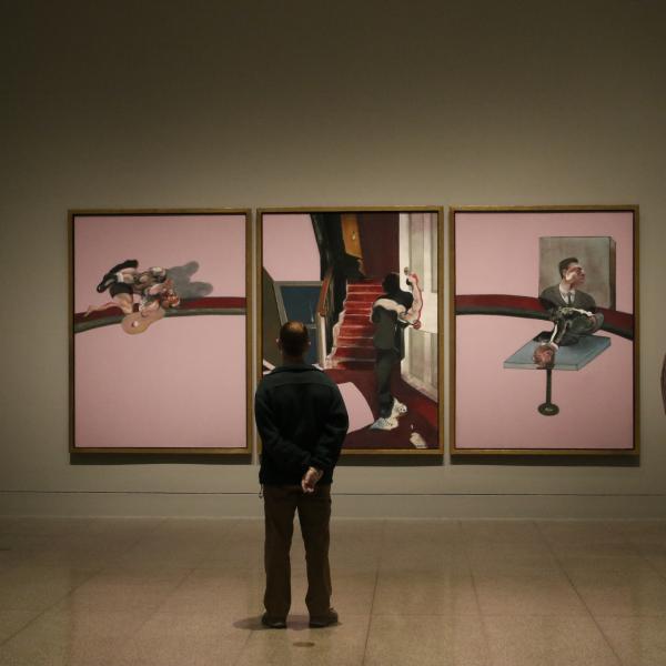 Masked museum visitors look at three large paintings by Francis Bacon