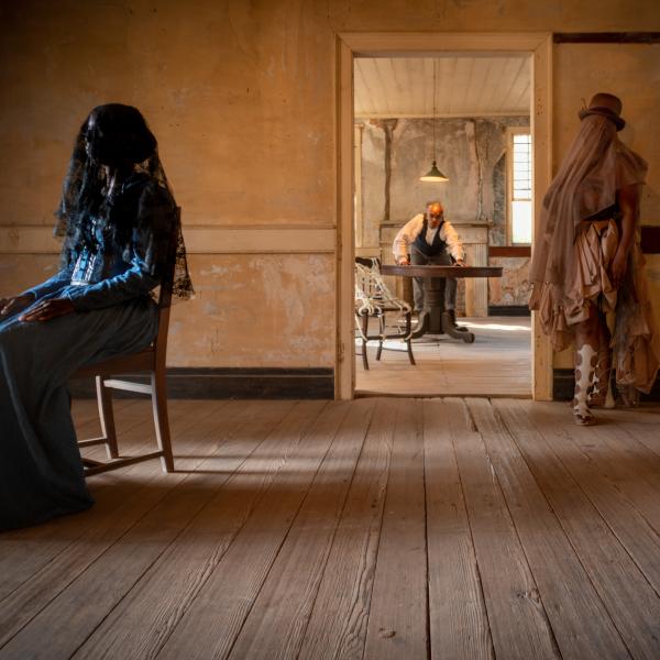 A Black woman in a blue dress and black veil sits in a chair to the left, a woman with a top hat and veil stands to the left and through the door way of a plantation mansion is a Black man standing over a table. 