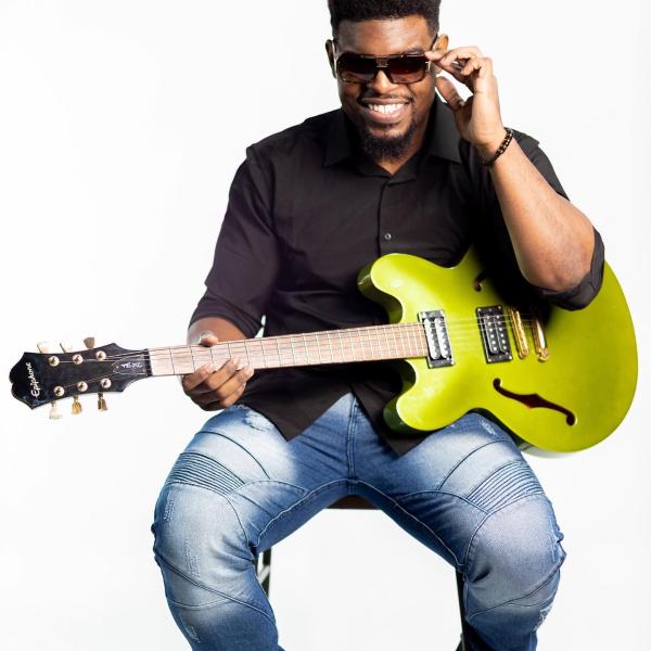 Man wearing sunglasses holding a guitar, sitting. 