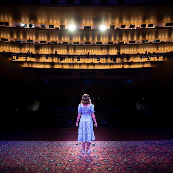 A woman in a blue dress stands on a stage facing a dark auditorium