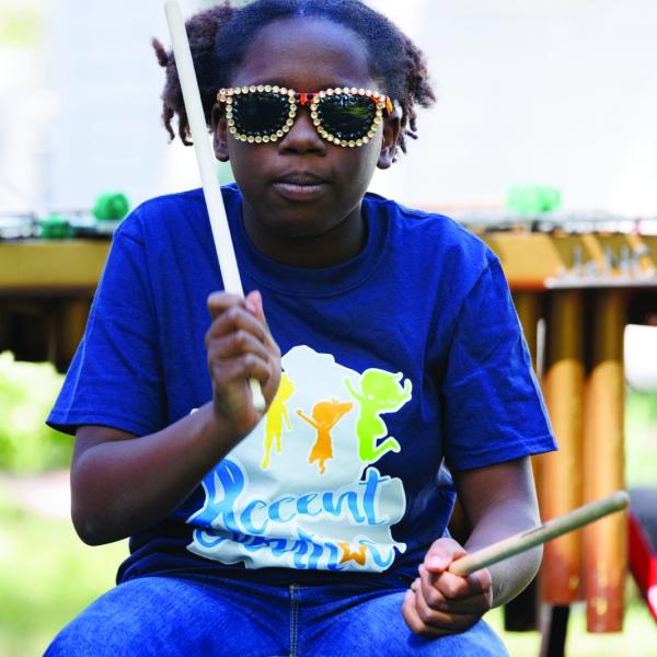 Girl wearing sunglasses and blue shirt drumming on stage outdoors. 
