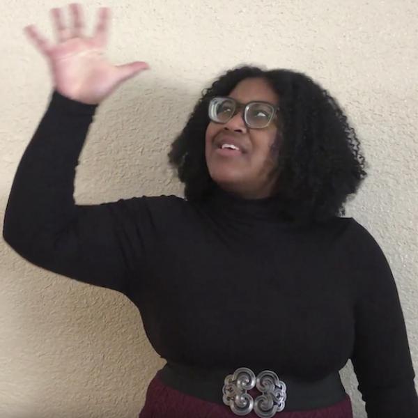 Woman in black turtleneck looks to sky and holds up hand with open palm towards camera 