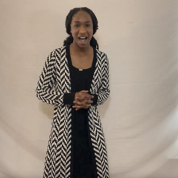 Woman in black dress speaks with fingers interlaced in front of her