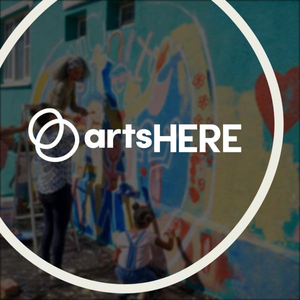 The artsHERE logo features two overlapping spheres. It’s framed by a circular outline. The logo is over a blurred image of people painting a colorful mural.
