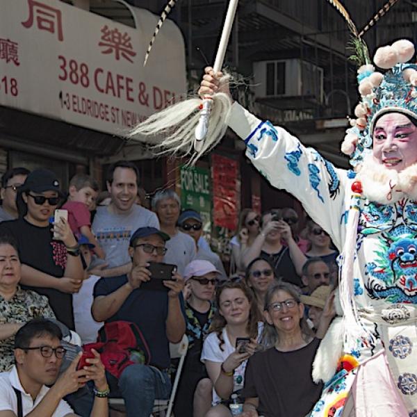 Elaborately costumed figure in parading down a city street surrounded by a large crowd