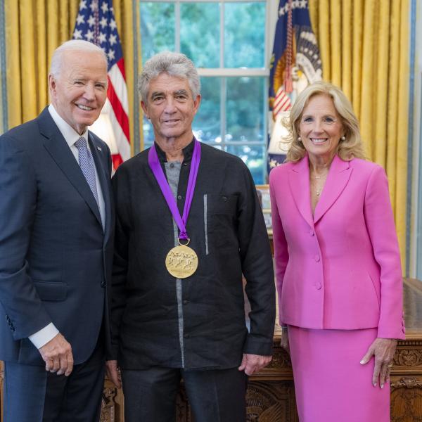 Man with gray hair wearing a medal around his neck surrounded by older man wearing a blue suit and woman with blonde hair wearing a pink suit. 