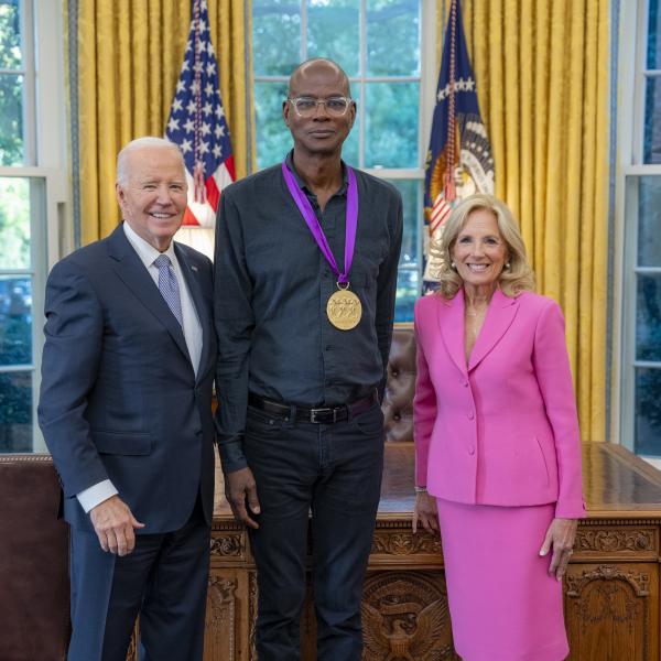 Tall bald Black man wearing glasses with a medal around his neck surrounded by older man wearing a blue suit and woman with blonde hair wearing a pink suit. 