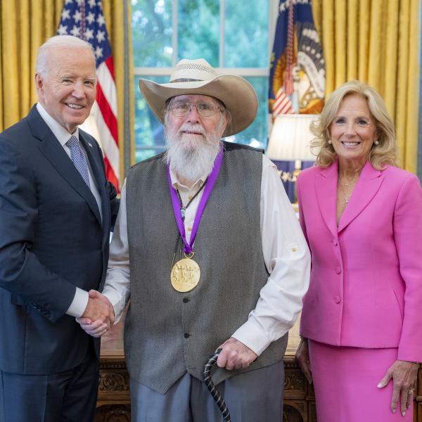 Man with cowboy hat and long gray beard wearing a medal around his neck surrounded by older man wearing a blue suit and woman with blonde hair wearing a pink suit. 