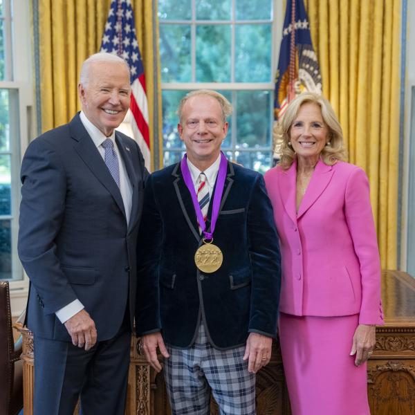 Man in blue velvet jacket wearing a medal around his neck surrounded by older man wearing a blue suit and woman with blonde hair wearing a pink suit. 