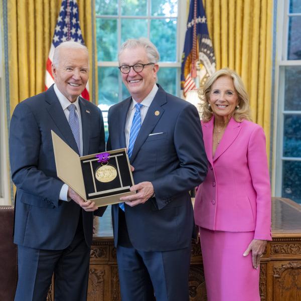 Man with gray hair and glasses holding a medal surrounded by older man wearing a blue suit and woman with blonde hair wearing a pink suit. 
