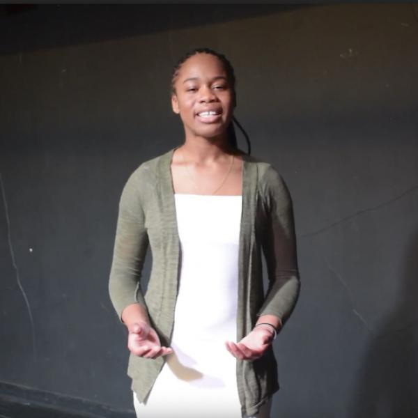 Woman in white dress and grey sweater looks at camera reciting