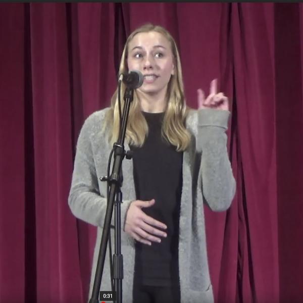 Woman in black shirt and grey sweater stands behind microphone reciting