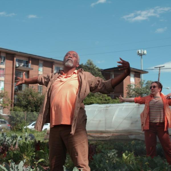 Black man with gray goatee wearing brown workshirt with arms extending in front of white woman in orange shirt doing same gesture, in a garden.