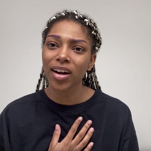Woman in black shirt recites to camera with hand on chest