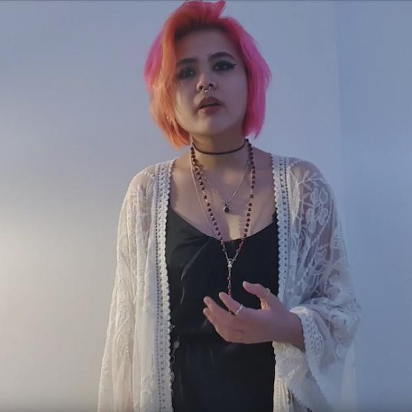 Woman in black dress and white jacket holds hand out as she recites towards camera