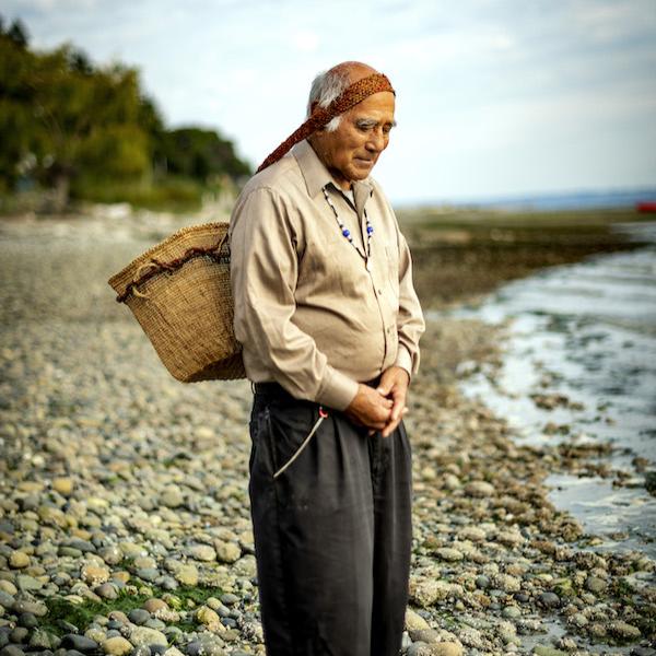 A man standing by the water with a basket strapped to his head.