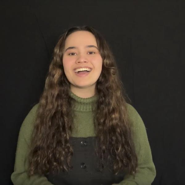 Woman in green turtleneck and black overalls recites to camera