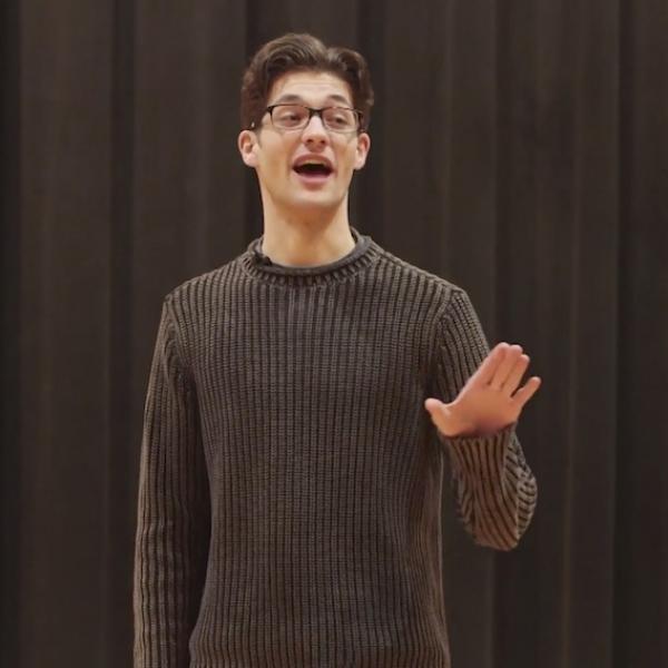 Man in grey sweater holds hand up with palm out as he recites on stage