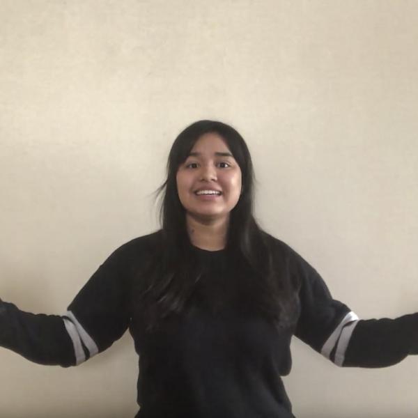 Woman in grey tshirt recites to camera with arms open and palms out