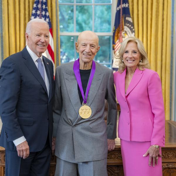 Older bald man in light blue suit wearing a medal around his neck surrounded by older man wearing a blue suit and woman with blonde hair wearing a pink suit. 