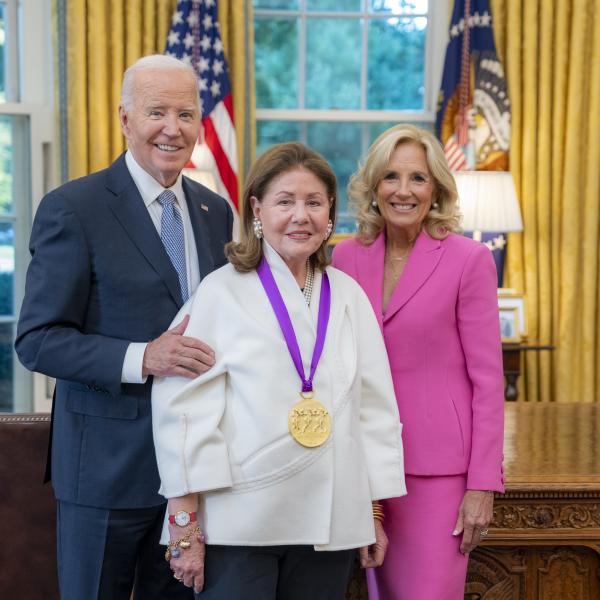 Woman with long brown hair in white shirt wearing a medal around her neck surrounded by older man wearing a blue suit and woman with blonde hair wearing a pink suit. 
