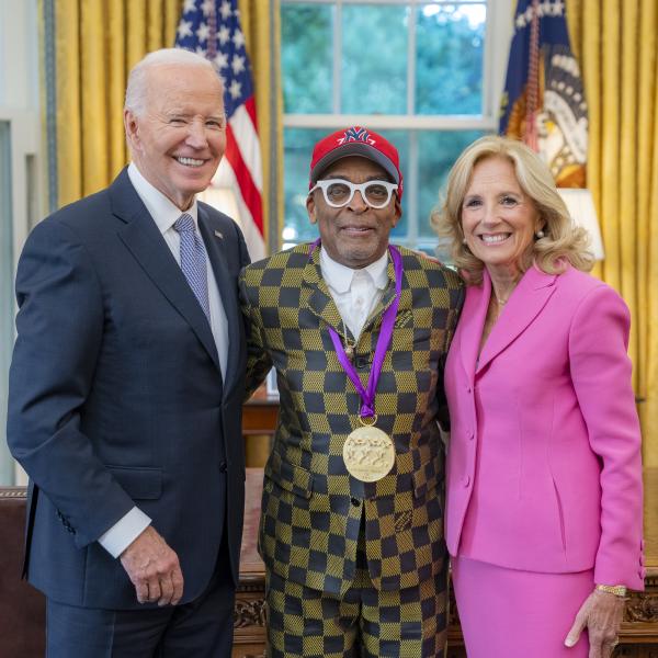 Black man with glasses and New York Yankees cap in a gold and blue checkered suit wearing a medal around his neck surrounded by older man wearing a blue suit and woman with blonde hair wearing a pink suit. 