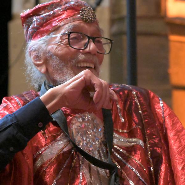 Black man with gray beard and glasses wearing red sequined suit, sitting, laughing. 