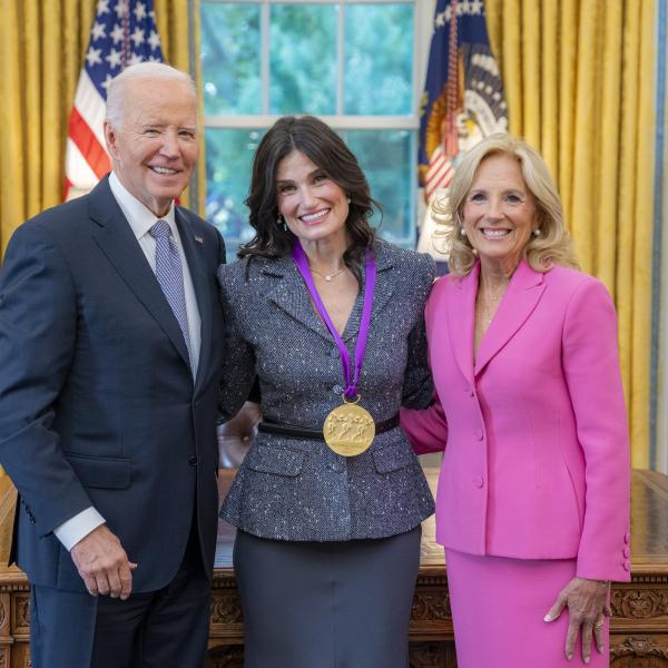 Woman with long dark hair in a gray suit wearing a medal around his neck surrounded by older man wearing a blue suit and woman with blonde hair wearing a pink suit. 