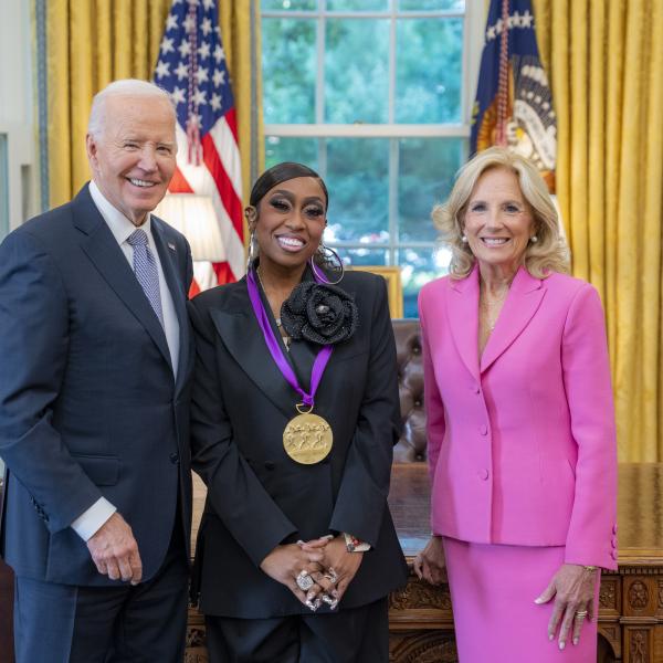 Black woman in dark blue suit wearing a medal around her neck surrounded by older man wearing a blue suit and woman with blonde hair wearing a pink suit. 